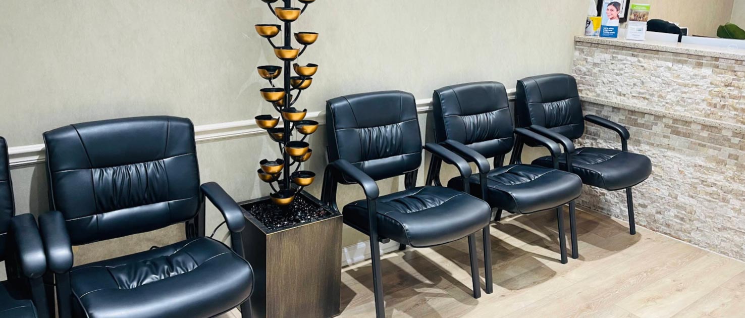 This is a photograph of an interior space featuring a row of black leather recliner chairs with armrests, arranged in front of a wall-mounted lamp with a yellow shade, and a decorative plant on top of a small table. The chairs are placed in a waiting area or reception room, which has a beige tiled floor and a light-colored wall.