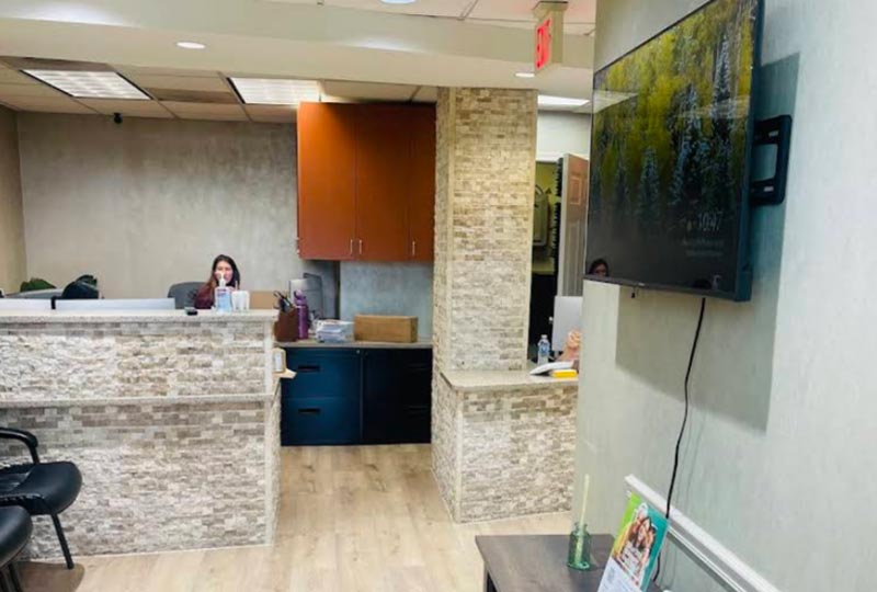 The image shows an interior view of a modern office space with a reception area featuring a desk with a computer monitor, a person seated at the desk, a flat-screen TV mounted on the wall displaying a program, a large window allowing natural light into the room, and a waiting area with a comfortable seating arrangement. The office has a professional ambiance with neutral colors and a clean design.