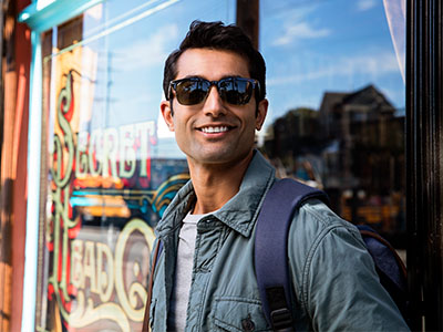 A young man wearing sunglasses and a backpack stands confidently on a city street, leaning against a storefront window with his hands in his pockets, smiling towards the camera.