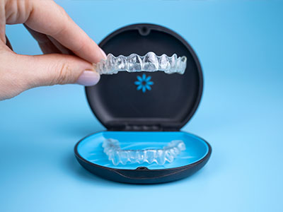 The image shows a person s hand holding an open dental retainer case containing a clear plastic aligner tray with multiple teeth-like structures, positioned on a blue surface against a light blue background.