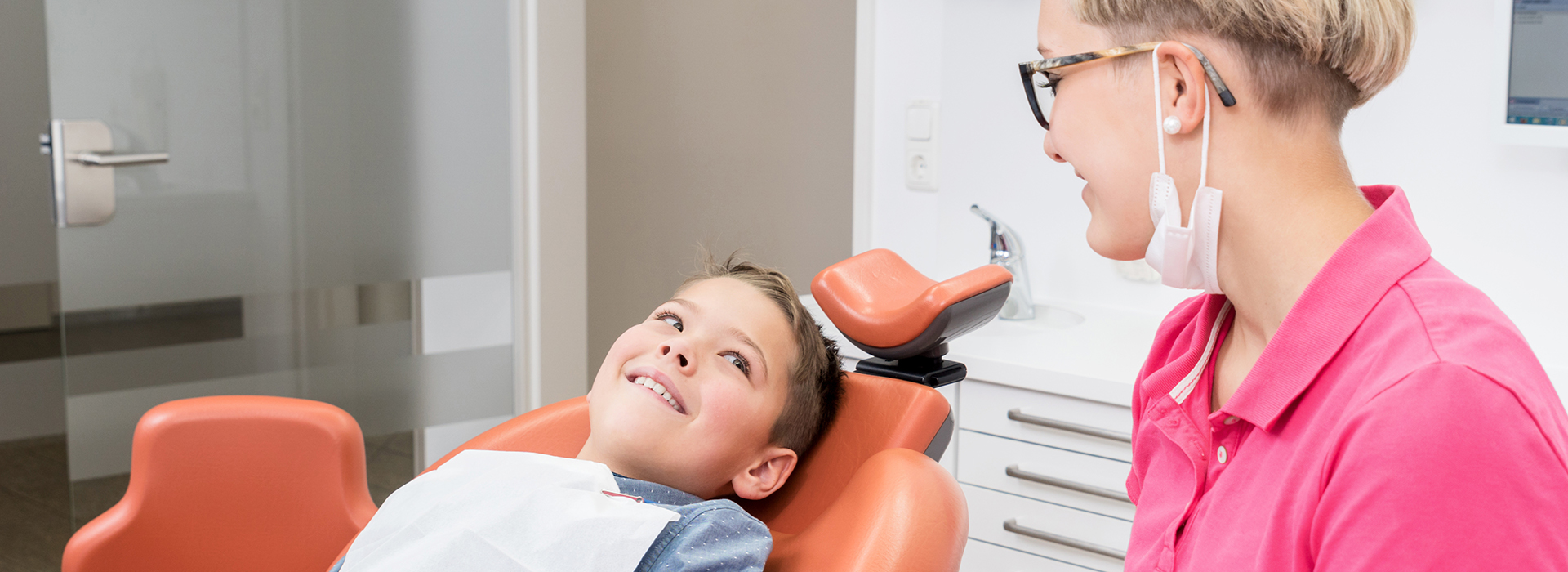 The image depicts a person sitting in a dental chair with a smiling face, being attended to by a dental professional who appears to be a woman, with both individuals wearing masks.