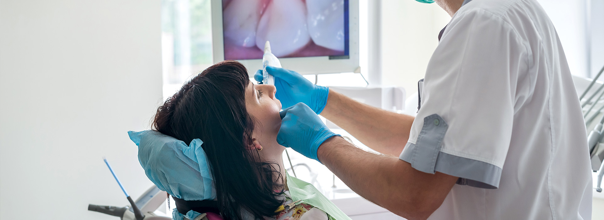 The image shows a dental professional performing a procedure on a patient s mouth using dental instruments, with the patient sitting in a chair and wearing a blue surgical mask.