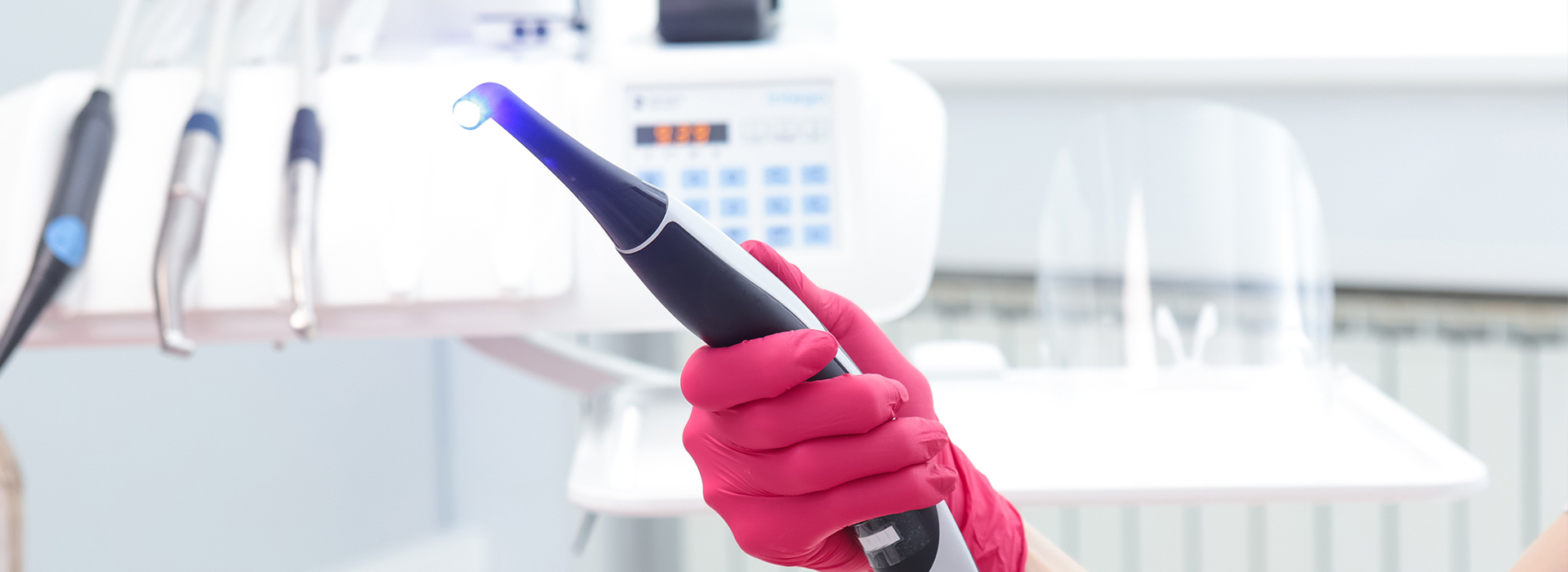 The image shows a person wearing red gloves holding an electronic device with a blue light, possibly a stethoscope, in front of a white wall with medical equipment and a blurred background that suggests a healthcare setting.