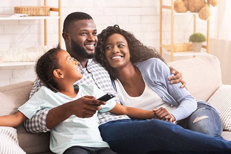 The image shows a man, woman, and child sitting together on a couch, with the man holding a remote control and smiling at the camera. They appear to be enjoying a family moment indoors.