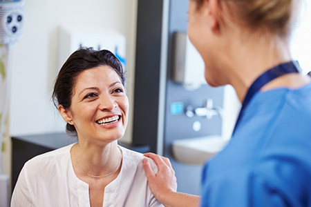 The image features two individuals in a medical setting, with a woman wearing scrubs smiling at another person who appears to be receiving care or attention.