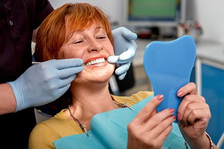 A woman with red hair is sitting in a dental chair, smiling at the camera while holding up a blue toothbrush.