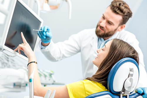 A woman sitting in a dental chair with a dentist standing beside her, both wearing face masks and gloves, examining a computer screen together in an office setting.
