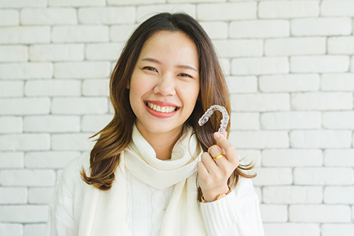 In the image, there is a woman with dark hair, wearing a white top and a scarf, holding up a toothbrush, smiling at the camera, standing against a brick wall background.