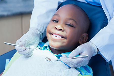 The image shows a young boy sitting in a dental chair, smiling at the camera, with a dental professional s hands visible near him, suggesting he is getting his teeth checked or cleaned.