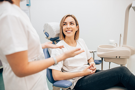 The image shows a woman sitting in a dental chair with her eyes closed, smiling, while being attended to by a dental professional who appears to be a dentist or hygienist.